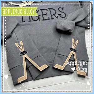 May include: Grey sweatshirt with the word "TIGERS" embroidered on the front. The sweatshirt has brown hockey stick appliques on the cuffs. The text "Applique Alley" is embroidered on the right cuff.