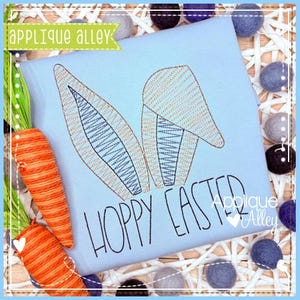Op de afbeelding: Een lichtblauw T-shirt met een gestikt konijnenoren-ontwerp en de tekst "Hoppy Easter" in zwart. Het shirt ligt op een witte achtergrond met decoratieve ballen.