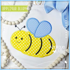 May include: White t-shirt featuring a yellow bee applique with white polka dots, black stripes, and blue polka dot wings. The bee has a smiling face and is embroidered with the text "Applique Alley". A blue bow and wooden beads are in the background.