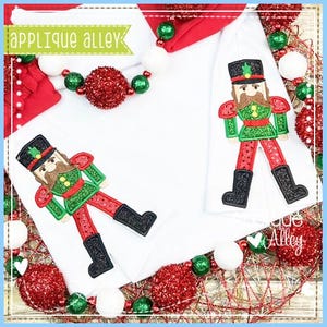 May include: White fabric with two embroidered nutcracker figures in green, red, black, and brown. The figures are surrounded by red and green Christmas ornaments and the text "applique alley".
