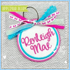 Puede incluir: Adorno redondo blanco con el nombre "Renleigh Mae" bordado en rosa. Tiene un borde rosa y turquesa, y está adornado con cintas rosas, blancas y turquesas. El adorno es de Applique Alley.