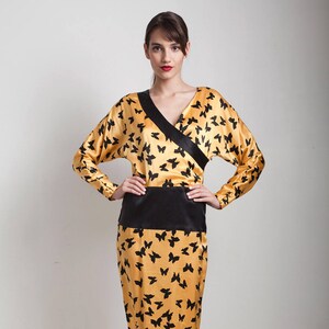 Puede incluir: Un vestido amarillo con estampado de mariposas negras y una cinta negra. El vestido tiene cuello en V y mangas largas.