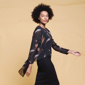 May include: A woman wearing a black skirt, black ankle boots, and a black top with a sheer overlay featuring a feather-like pattern in shades of brown, orange, and white. The background is a solid light yellow.