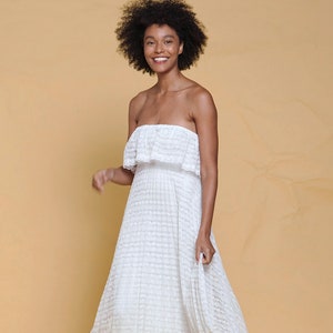 Puede incluir: Una mujer con un vestido blanco de tirantes sin tirantes y un patr&oacute;n de encaje. Ella est&aacute; sonriendo y caminando hacia la c&aacute;mara.