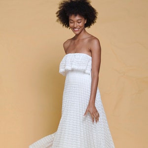 Puede incluir: Un vestido blanco maxi sin tirantes con una superposici&oacute;n de encaje y un escote con volantes. El vestido tiene una falda fluida y se lleva con zapatillas blancas.