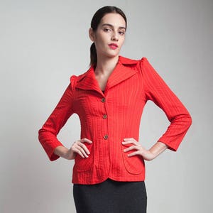 May include: A woman wearing a red blazer with a black skirt and brown sandals. The blazer has a collar and buttons down the front.