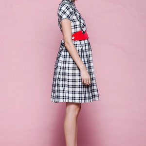 May include: A short-sleeved plaid dress in black and white, cinched at the waist with a bright red belt. The dress is paired with red wedge heels. The background is a solid pink.