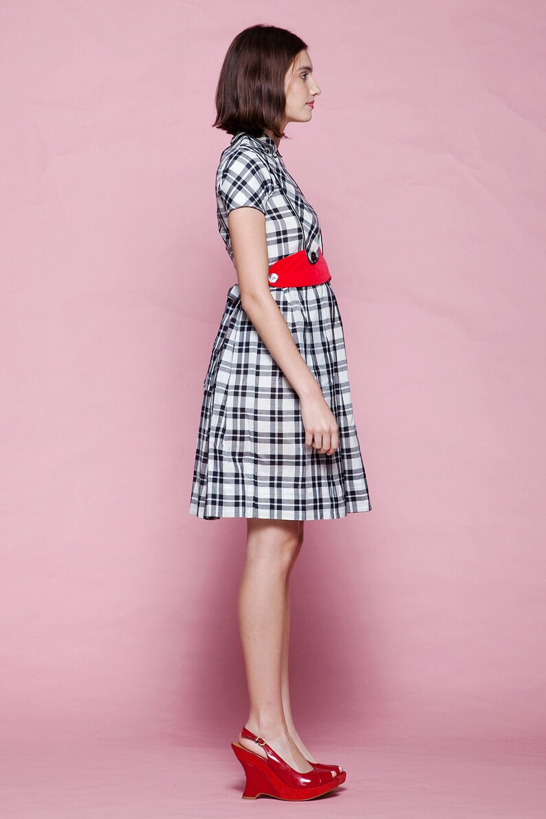 May include: A short-sleeved plaid dress in black and white, cinched at the waist with a bright red belt. The dress is paired with red wedge heels. The background is a solid pink.