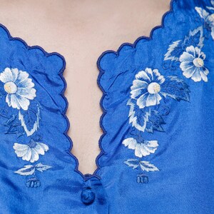 Puede incluir: Una blusa de sat&eacute;n azul con un escote festoneado y flores bordadas blancas.