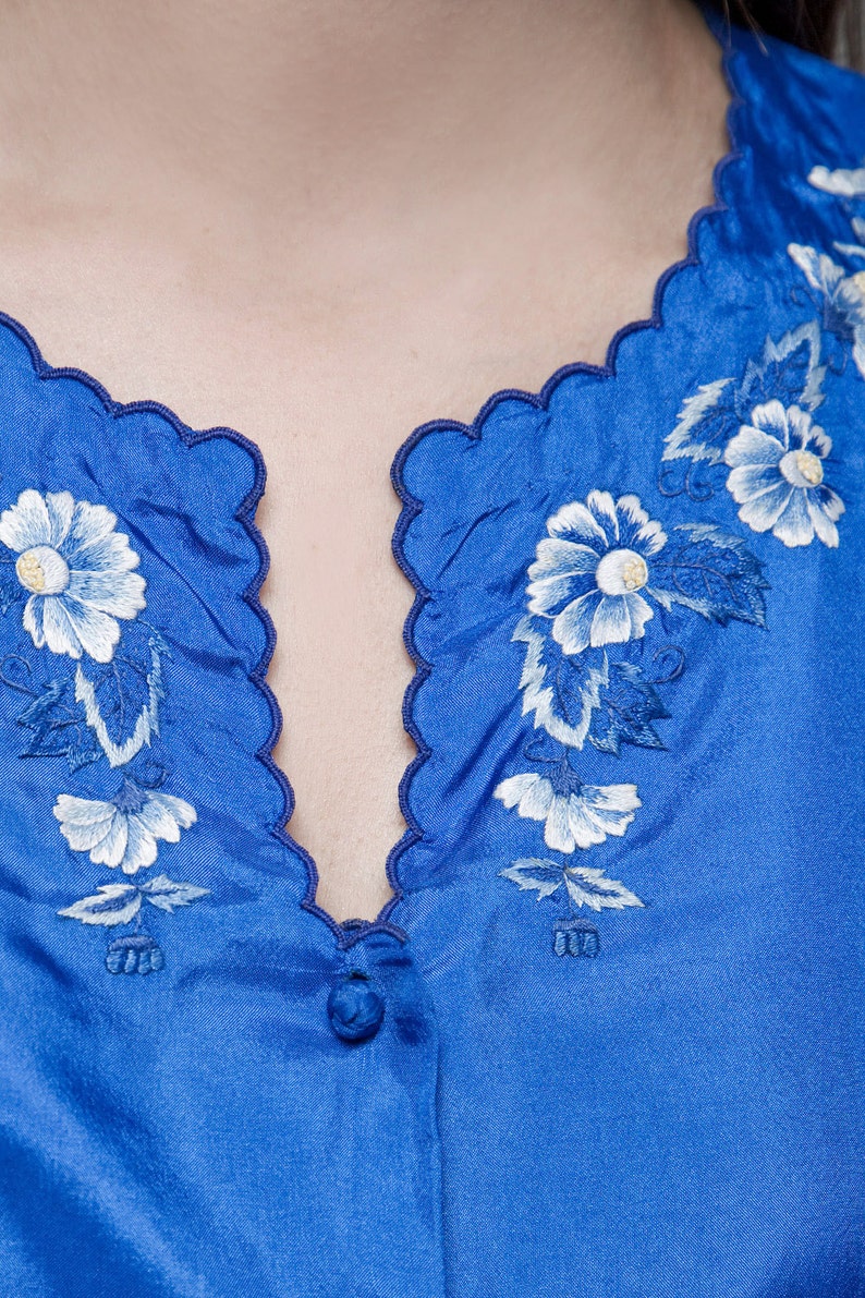 Puede incluir: Una blusa de sat&eacute;n azul con un escote festoneado y flores bordadas blancas.