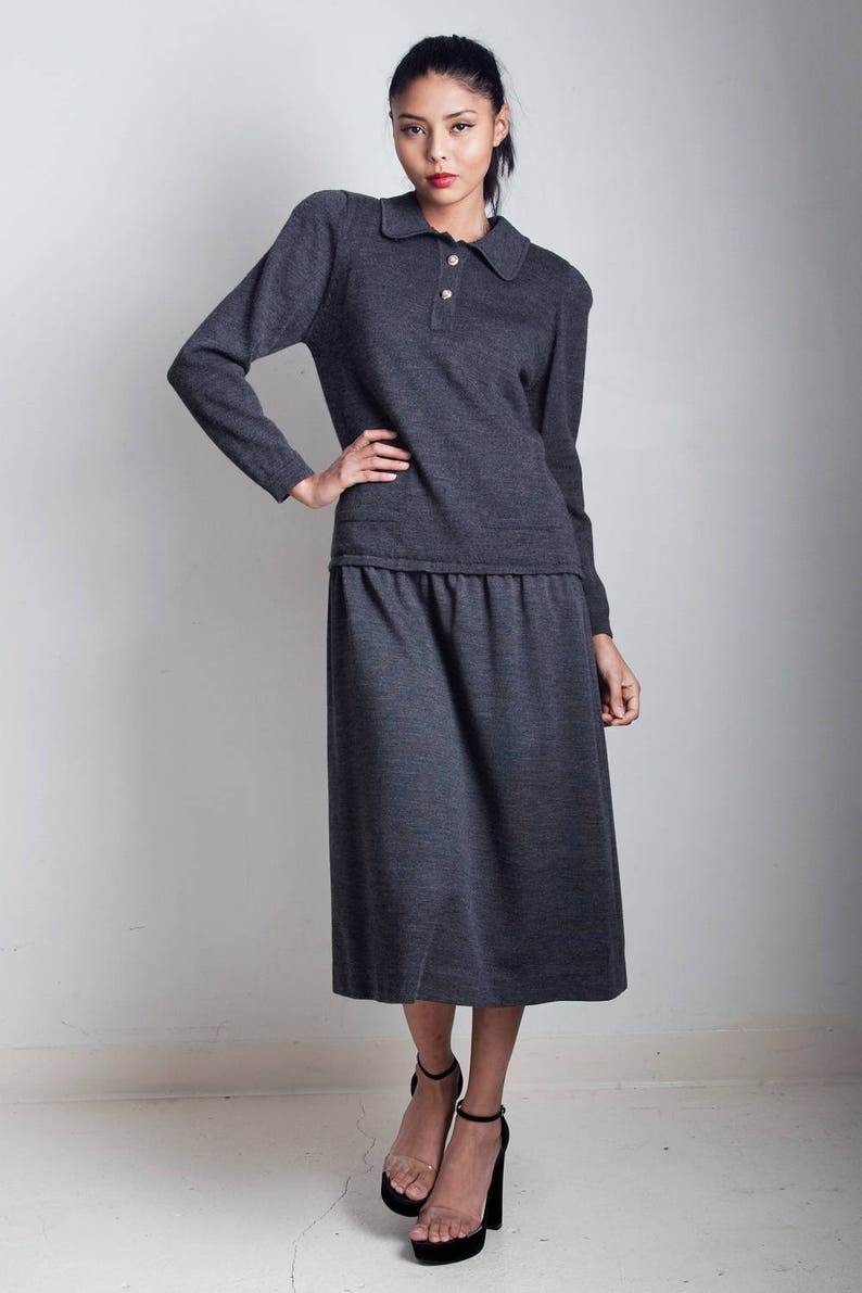 May include: A charcoal gray knit sweater dress with a collared neckline and button closure. The dress has a fitted bodice and a slightly flared skirt. The model is wearing black heeled sandals.