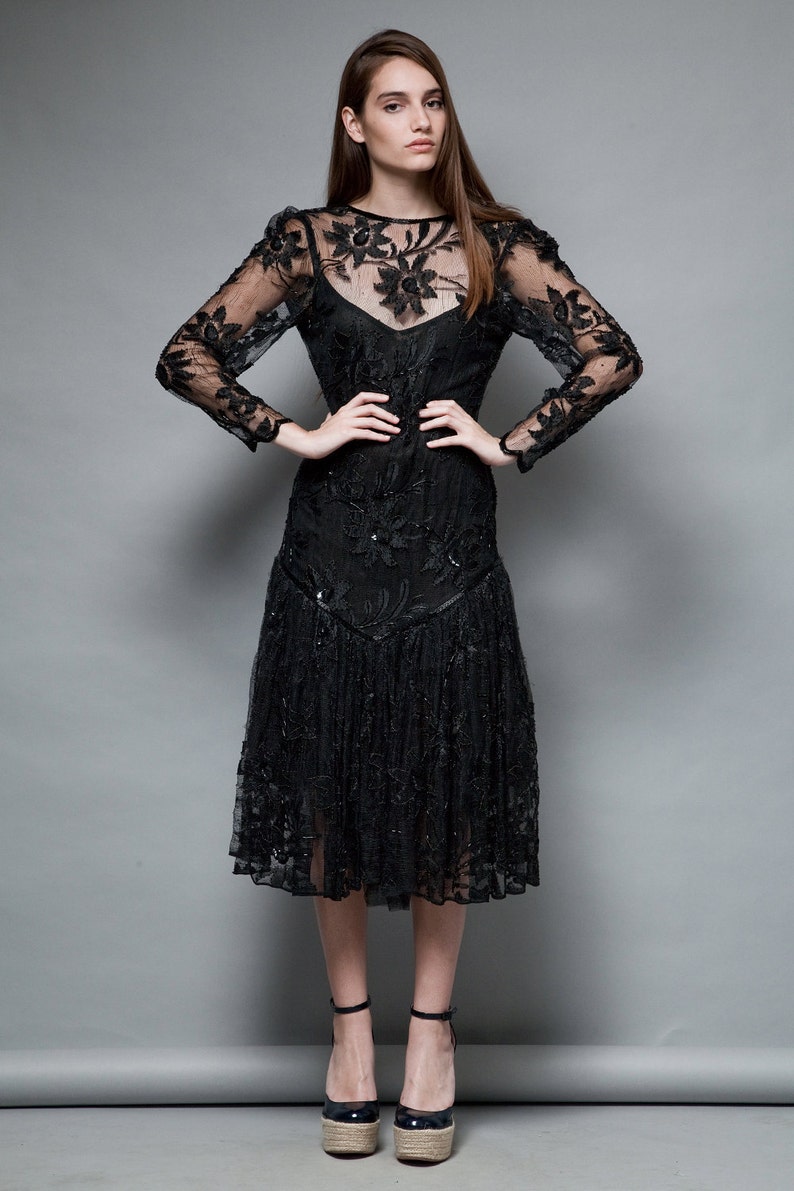May include: A black lace dress with a floral pattern and a ruffled hem. The dress has long sleeves and a fitted bodice. The model is wearing the dress with black heeled shoes.