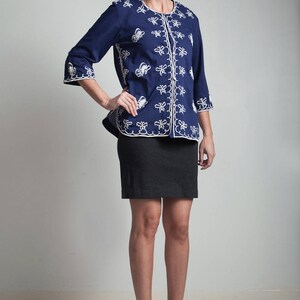 May include: A navy blue embroidered jacket with white floral designs. The jacket has a button-up front and three-quarter sleeves. The model is wearing a black skirt and brown platform heels.