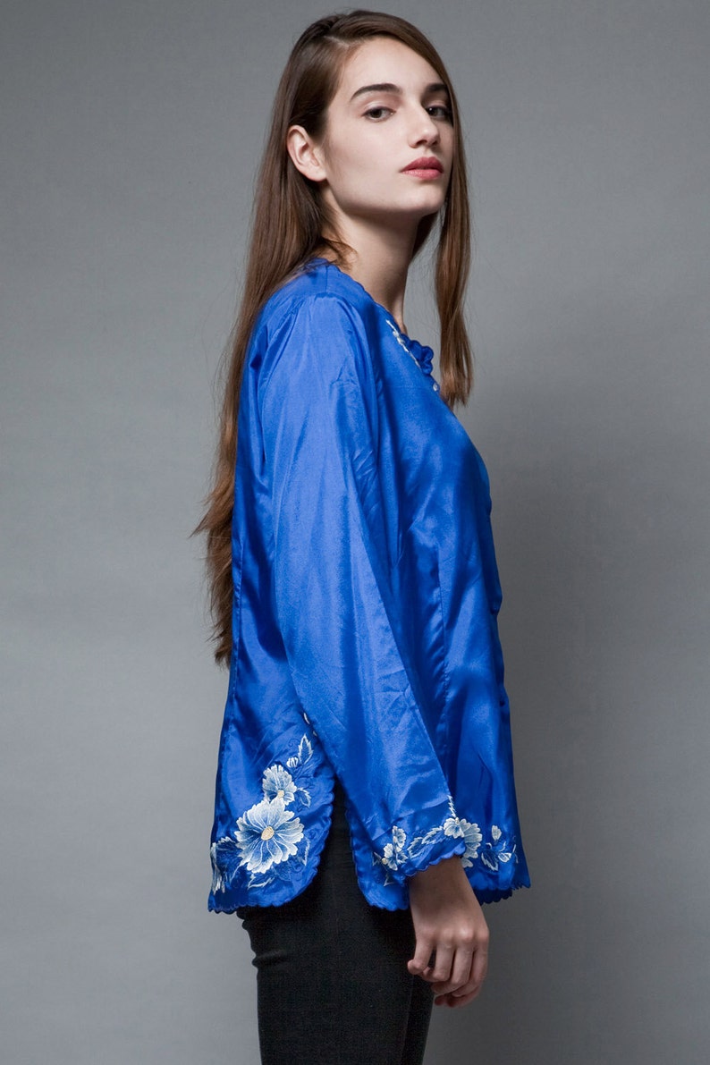 Puede incluir: Una blusa de sat&eacute;n azul con bordados florales blancos en las mangas y el dobladillo.