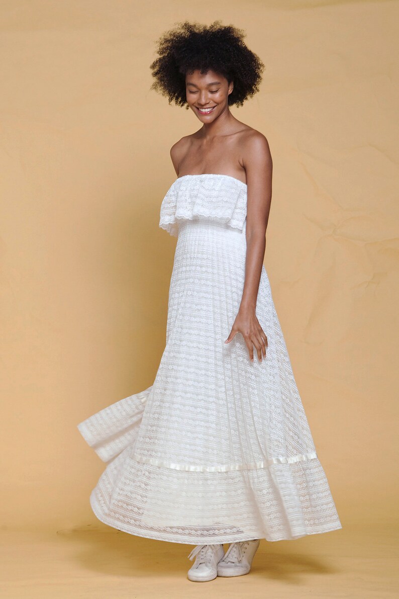 Puede incluir: Un vestido blanco maxi sin tirantes con una superposici&oacute;n de encaje y un escote con volantes. El vestido tiene una falda fluida y se lleva con zapatillas blancas.