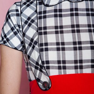 May include: A vintage-style dress featuring a black and white plaid pattern. The dress has a contrasting red belt and a rounded collar. The design includes black piping and a button detail. The dress is a classic, retro garment.