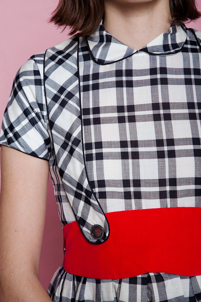 May include: A vintage-style dress featuring a black and white plaid pattern. The dress has a contrasting red belt and a rounded collar. The design includes black piping and a button detail. The dress is a classic, retro garment.