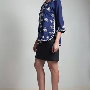 May include: A woman wearing a navy blue embroidered jacket with white stitching and a black skirt. She is wearing brown heeled sandals with white soles.