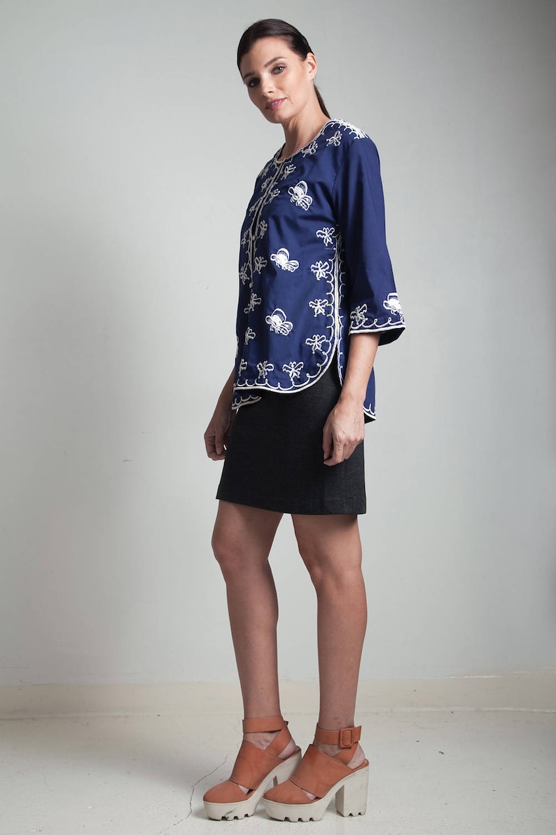 May include: A woman wearing a navy blue embroidered jacket with white stitching and a black skirt. She is wearing brown heeled sandals with white soles.