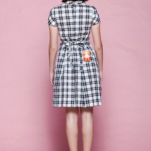 May include: A short-sleeved plaid dress in black and white, featuring a fitted bodice and a pleated skirt. The dress has a tie-back detail at the waist and is paired with red heels. A small tag is attached to the dress. The background is a solid pink.