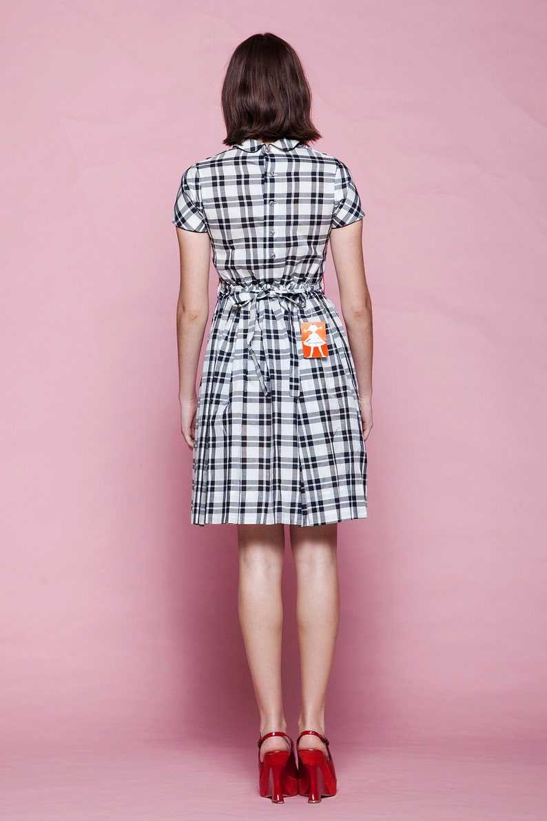 May include: A short-sleeved plaid dress in black and white, featuring a fitted bodice and a pleated skirt. The dress has a tie-back detail at the waist and is paired with red heels. A small tag is attached to the dress. The background is a solid pink.