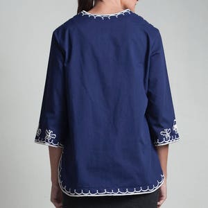 May include: A navy blue, 3/4 sleeve blouse with white embroidered trim around the neckline and cuffs. The blouse has a relaxed fit and is paired with a dark gray skirt.