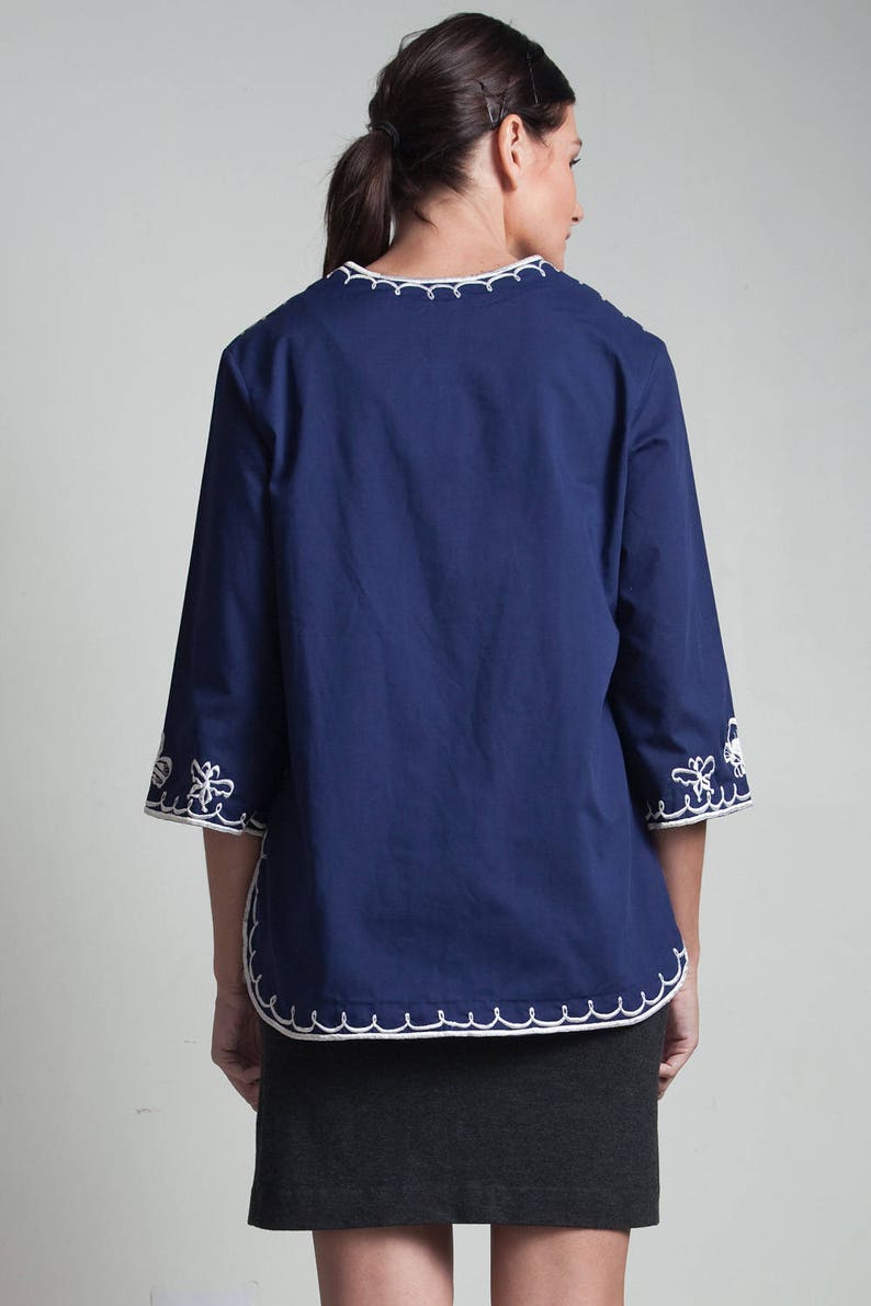 May include: A navy blue, 3/4 sleeve blouse with white embroidered trim around the neckline and cuffs. The blouse has a relaxed fit and is paired with a dark gray skirt.