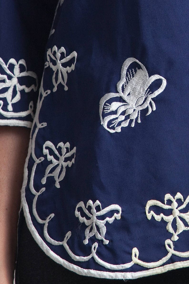 May include: A navy blue fabric with white embroidered butterflies and floral designs. The fabric has a scalloped edge.