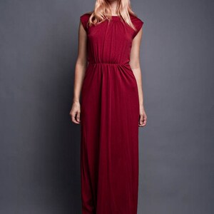Pu&ograve; includere: Una donna indossa un lungo abito maxi color borgogna con scollo rotondo e maniche corte. L'abito ha un corpetto aderente e una gonna fluida. Porta i capelli biondi lisci con la frangia.
