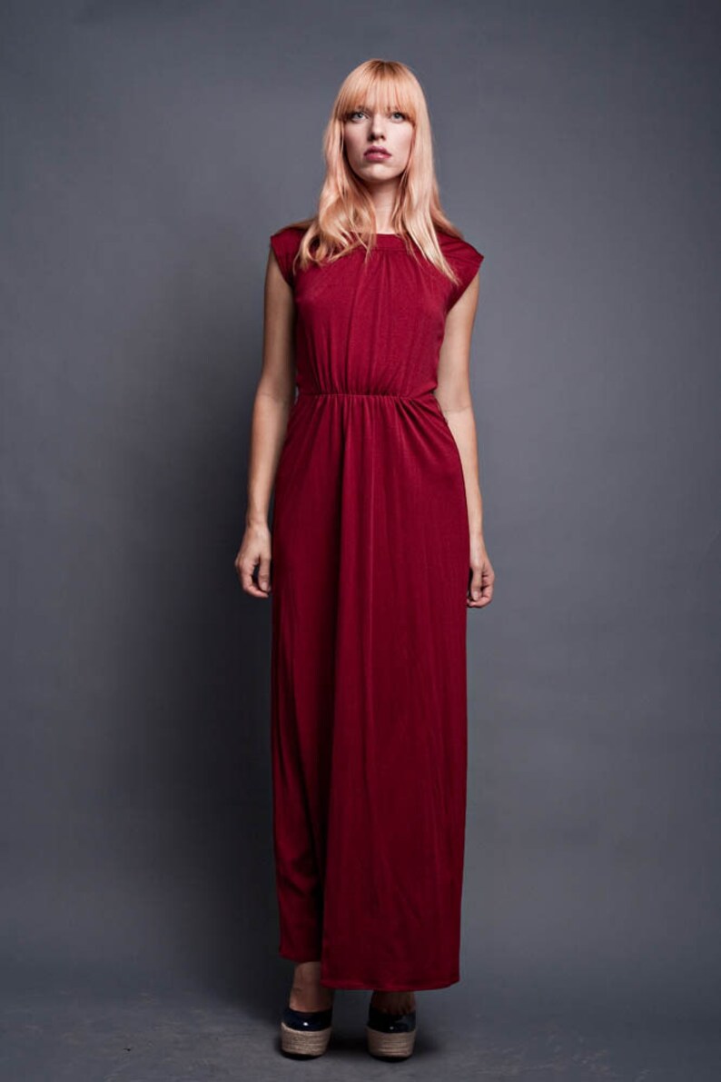 Pu&ograve; includere: Una donna indossa un lungo abito maxi color borgogna con scollo rotondo e maniche corte. L'abito ha un corpetto aderente e una gonna fluida. Porta i capelli biondi lisci con la frangia.