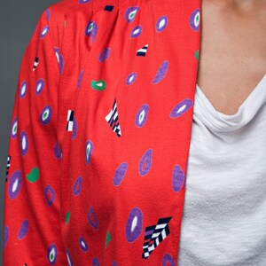 May include: A red jacket with a geometric pattern of purple, green, and black shapes. The jacket has a collar and a button closure.