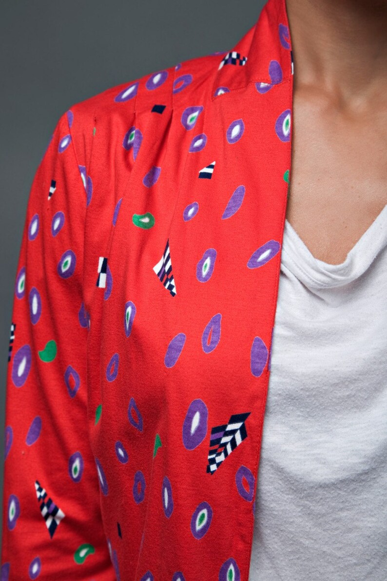 May include: A red jacket with a geometric pattern of purple, green, and black shapes. The jacket has a collar and a button closure.