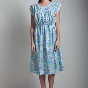 May include: A woman wearing a light blue dress with a floral print and white lace trim. The dress has a cinched waist and a flowing skirt. She is wearing beige heeled sandals.
