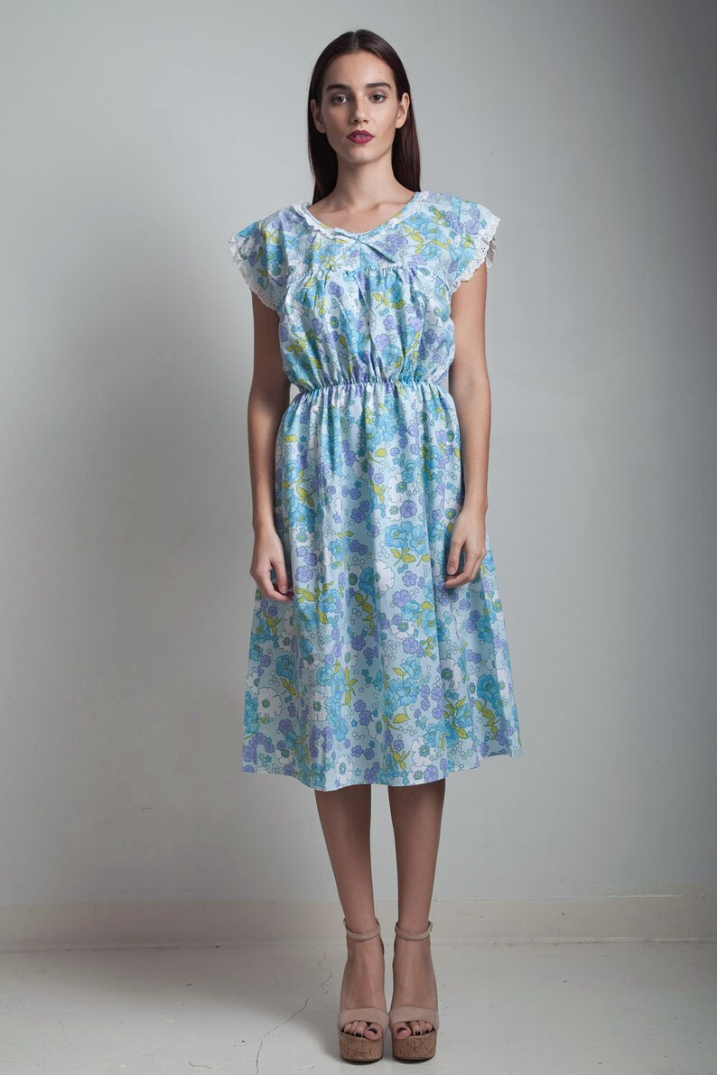 May include: A woman wearing a light blue dress with a floral print and white lace trim. The dress has a cinched waist and a flowing skirt. She is wearing beige heeled sandals.