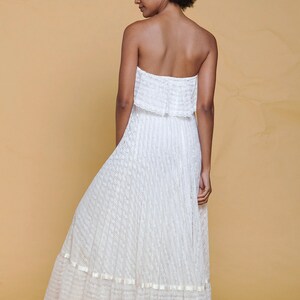 Puede incluir: Un vestido blanco maxi sin tirantes con una superposici&oacute;n de encaje y un dobladillo con volantes. El vestido est&aacute; hecho de una tela ligera y tiene una silueta fluida.