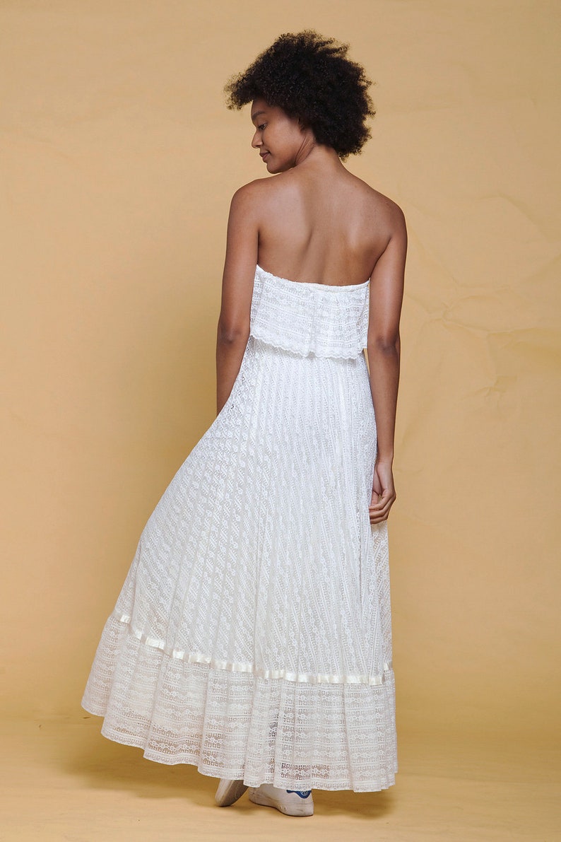 Puede incluir: Un vestido blanco maxi sin tirantes con una superposici&oacute;n de encaje y un dobladillo con volantes. El vestido est&aacute; hecho de una tela ligera y tiene una silueta fluida.
