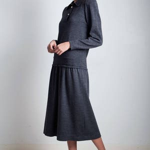 May include: A charcoal gray long-sleeved dress with a collared neckline and a gathered waist. The dress is made of a soft, knit fabric and has a relaxed fit. The model is wearing black heeled sandals.