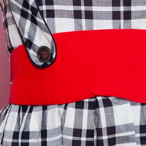 May include: A black and white plaid dress with a wide red belt. The dress features a button detail on the bodice and a flared skirt. The fabric has a classic gingham pattern.