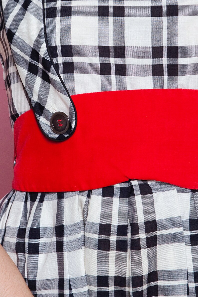 May include: A black and white plaid dress with a wide red belt. The dress features a button detail on the bodice and a flared skirt. The fabric has a classic gingham pattern.