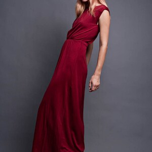 Pu&ograve; includere: Una donna indossa un lungo abito maxi color borgogna con un design senza maniche. &Egrave; in piedi su uno sfondo grigio e indossa zeppe.
