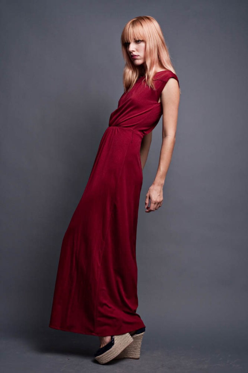 Pu&ograve; includere: Una donna indossa un lungo abito maxi color borgogna con un design senza maniche. &Egrave; in piedi su uno sfondo grigio e indossa zeppe.