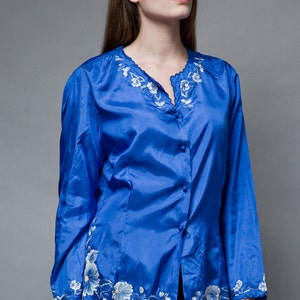 Puede incluir: Una blusa de sat&eacute;n azul real con un dise&ntilde;o de bordado floral blanco alrededor del cuello y el dobladillo. La blusa tiene un frente abotonado y mangas largas.
