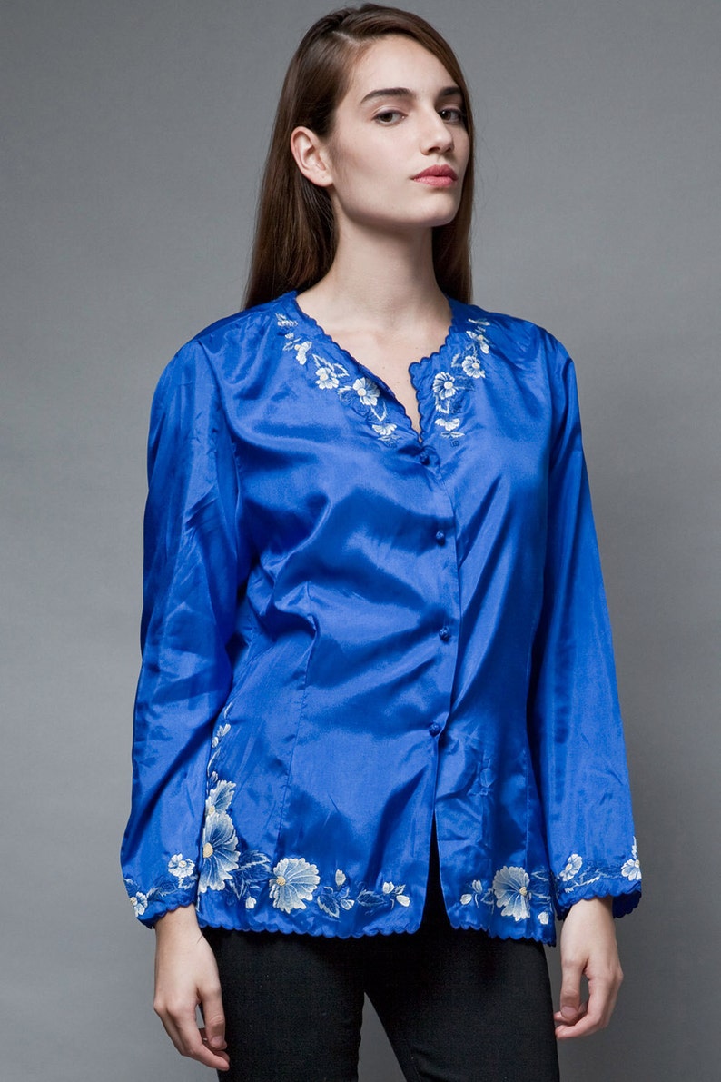 Puede incluir: Una blusa de sat&eacute;n azul real con un dise&ntilde;o de bordado floral blanco alrededor del cuello y el dobladillo. La blusa tiene un frente abotonado y mangas largas.