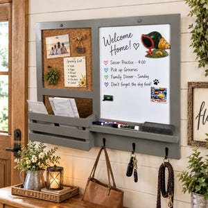 May include: A gray wall-mounted organizer featuring a cork bulletin board, a white dry-erase board with the text Welcome Home, a mail slot, and three black hooks. The board displays a grocery list, a photo, and a seashell magnet.