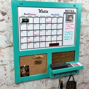 May include: A turquoise-framed magnetic dry erase calendar with a corkboard and key hooks. The calendar displays the month of February 2023, with notes and a small photo. Includes a shelf for markers and an eraser. The text "Watts" is at the top.