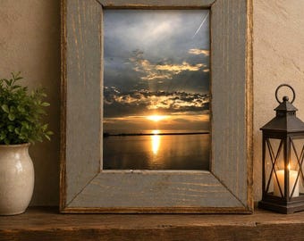 May include: A rectangular picture frame with a weathered gray finish and gold-toned edges, containing a photograph of a sunset over a body of water with dark clouds and a bright horizon line reflecting on the surface of the calm water below.