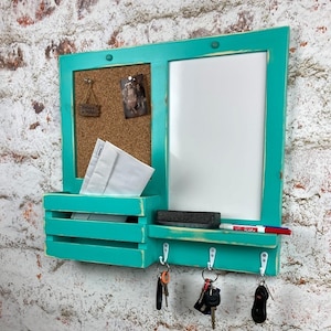 May include: Teal blue wooden wall organizer with a corkboard, a white dry erase board, a shelf with hooks, and a small mail slot. The corkboard has a small picture and a sign that says "Believe in Magic".
