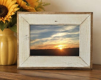 May include: A rectangular picture frame with a distressed white finish and visible wood grain texture. The frame contains a photograph of a sunset over a dark horizon with clouds in shades of orange, yellow, and blue across the sky.