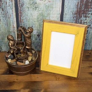 May include: A small tabletop fountain with two figures of children, one holding a bucket and the other holding a rope, surrounded by small stones. The fountain is in a wooden bucket and is sitting on a wooden surface. A yellow picture frame is to the right of the fountain.