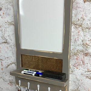 May include: Gray wooden wall organizer with a white dry erase board, a shelf for storing markers and a sponge, and a row of hooks for hanging keys and bags.
